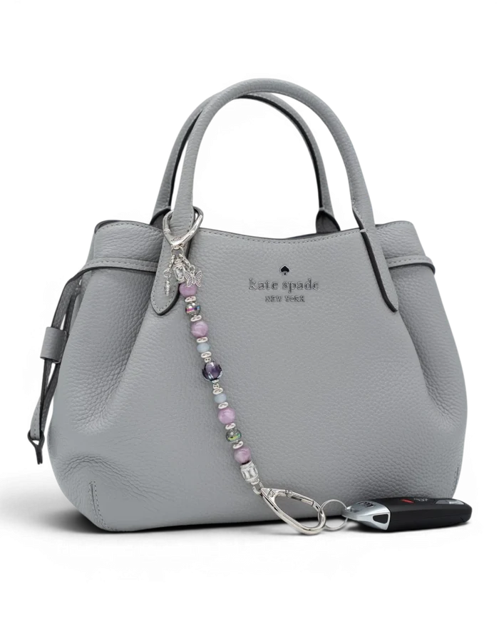 LD Keyfinder Blooming Joy beaded accessory with lavender lepidolite and glass charms attached to a grey leather handbag