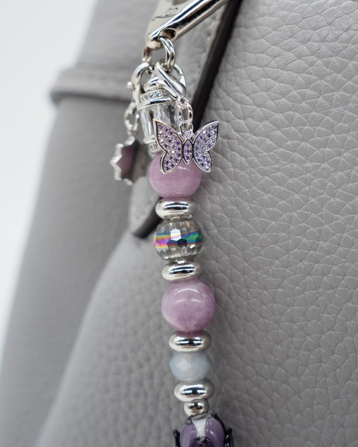 LD Keyfinder Blooming Joy accessory with lavender Lepidolite, purple Jade, and a glass tulip charm on a grey handbag.