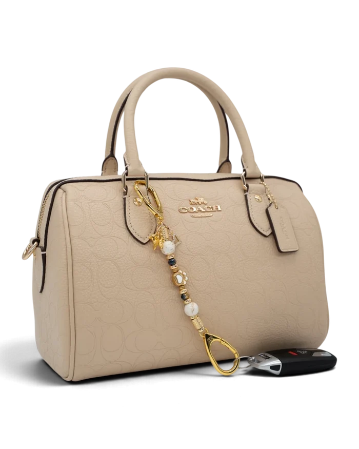 LD Keyfinder Furever Love-Mini attached to a beige Coach handbag, featuring white seashell and gold-plated dog charms.