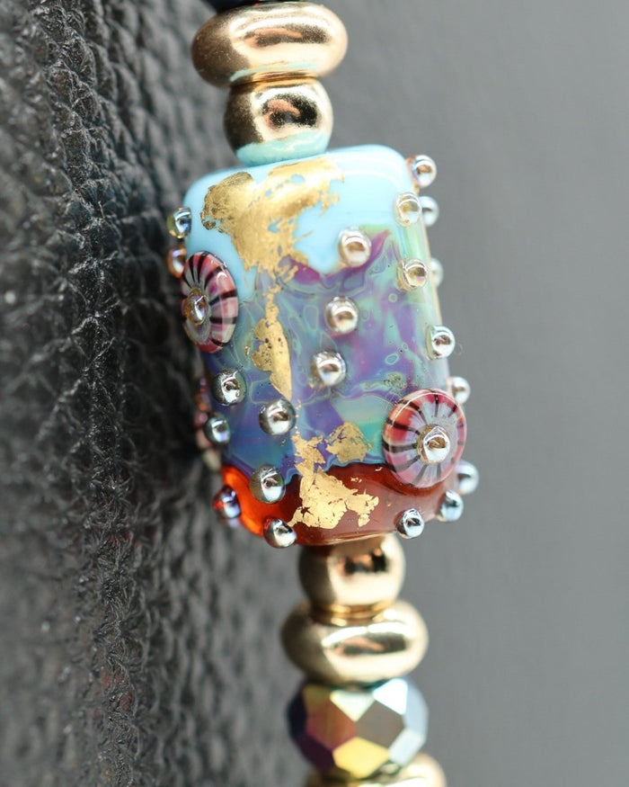 LD Keyfinder Gilded Fantasy bracelet with a central handcrafted glass bead featuring sky blue, topaz, 23KT gold leaf, and Murrini. Accented with gold-plated beads and blue flower charm.