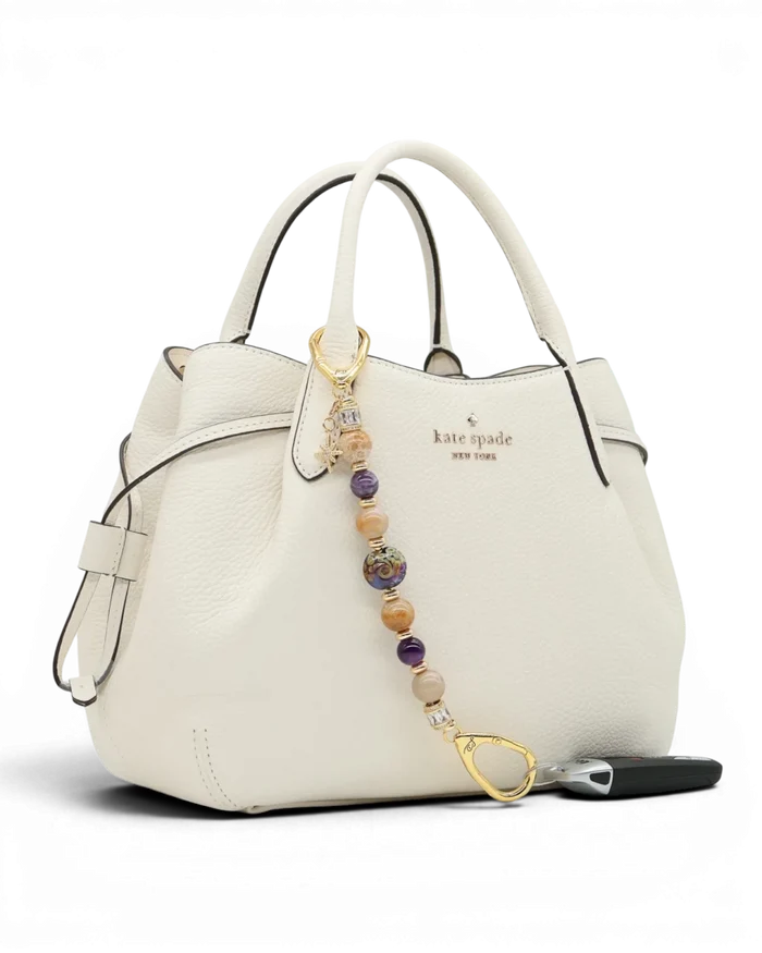 LD Keyfinder Golden Nebula charm with purple, gold, and amethyst beads, gold-plated star charm on white handbag.