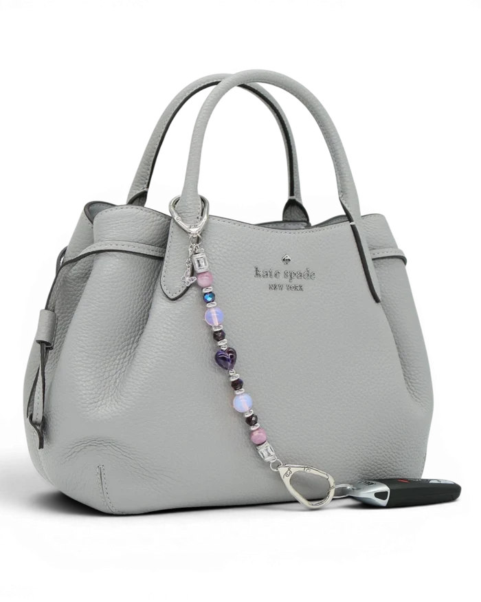 LD Keyfinder Orchid Flutter beaded accessory with lepidolite, opalite, and butterfly charm attached to a grey handbag.