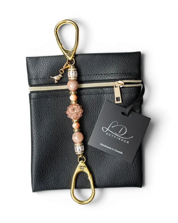 LD Keyfinder Petite Birdsong Bliss key chain with peach glass, sunstone beads, and gold bird charm on a black pouch.
