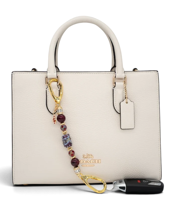 LD Keyfinder Petite Bordeaux Bloom accessory with burgundy agate, artisan glass, and gold charms on a white handbag.